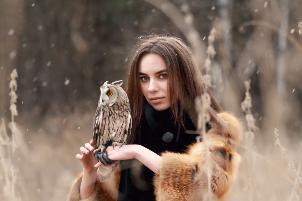 Woman in fur coat with owl on hand by first autumn snow. Beautiful brunette with long hair in nature