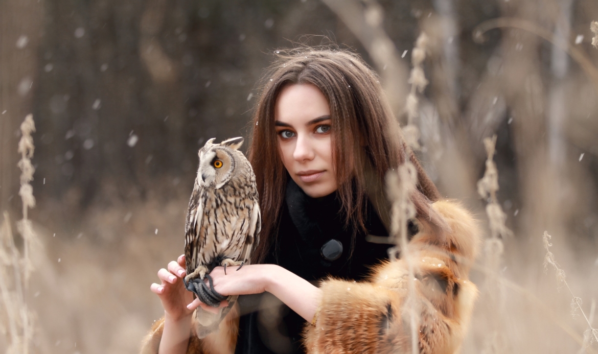 Woman in fur coat with owl on hand by first autumn snow. Beautiful brunette with long hair in nature