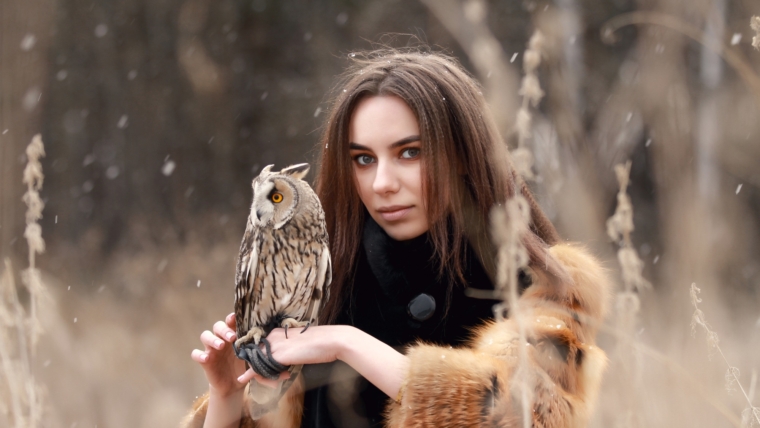 Woman in fur coat with owl on hand by first autumn snow. Beautiful brunette with long hair in nature